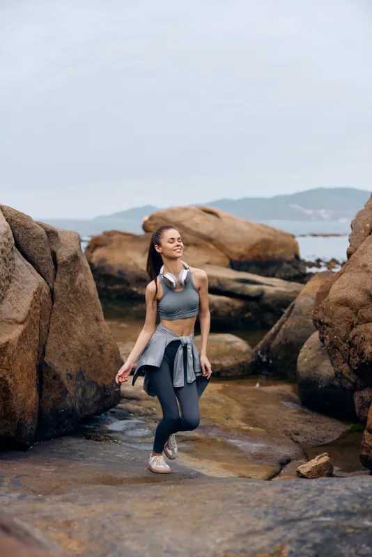 Young athletic woman exercising outdoors, wearing gray sportswear, amidst rocky terrain, embodying strength and vitality in a natural coastal setting