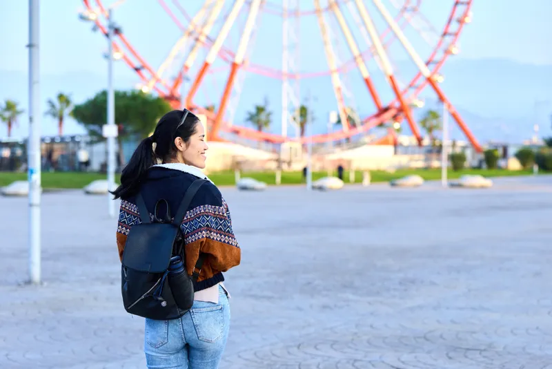 Woman with backpack walking toward a colorful ferris wheel at an outdoor amusement park, exploring grounds, casual style, daytime scene, cheerful mood, travel vibe, light breeze, distant palm trees
