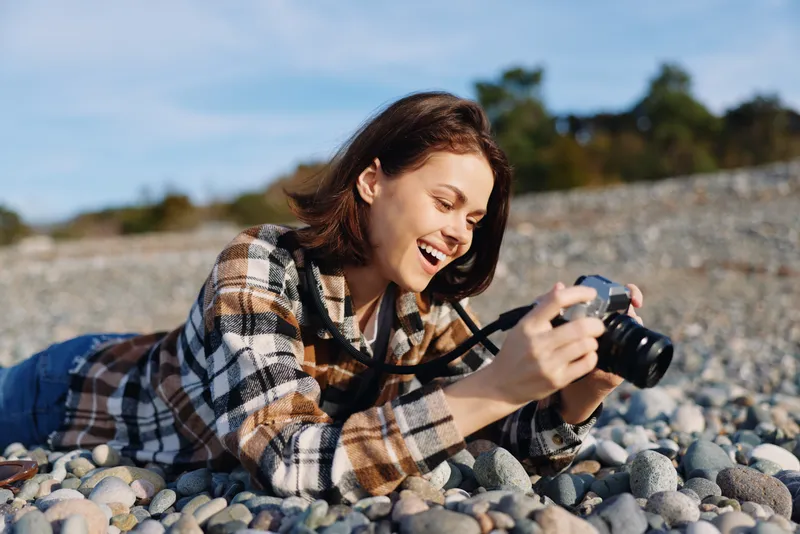 Woman photographer on a pebble beach captures a joyful moment with a camera, lying on stones in a casual plaid shirt under a bright sky during sunny day
