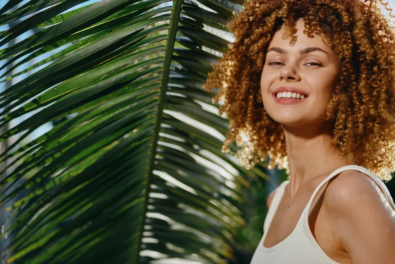 smiling woman with curly hair stands near tropical palm leaves outdoors, wearing a white tank top in bright natural light, expressing happiness and warmth
