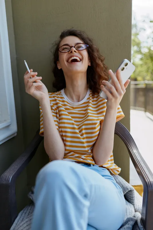 Smiling woman with curly hair wearing a striped tee relaxing on a balcony, holding a cigarette in one hand and a smartphone in the other. Casual home lifestyle, cheerful mood, and relaxed routine.