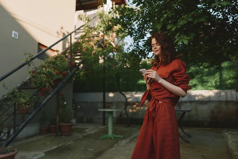 A woman in a red dress stands outdoors in a garden, holding a cup of coffee, smiling in a casual candid moment. Bright natural light highlights relaxed, everyday lifestyle, warmth, and charm