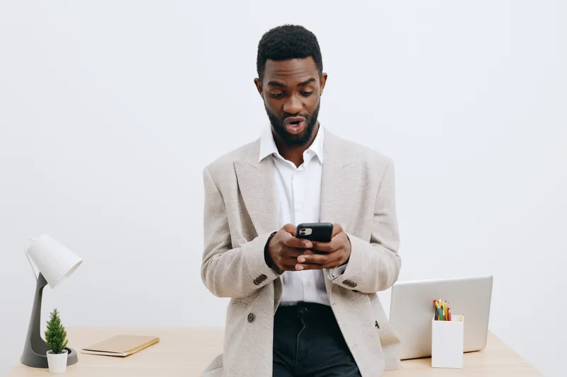 Modern African American man in beige blazer and white shirt using smartphone while standing at minimalist desk with laptop, pen holder, and plant inside contemporary office space. Business and technol