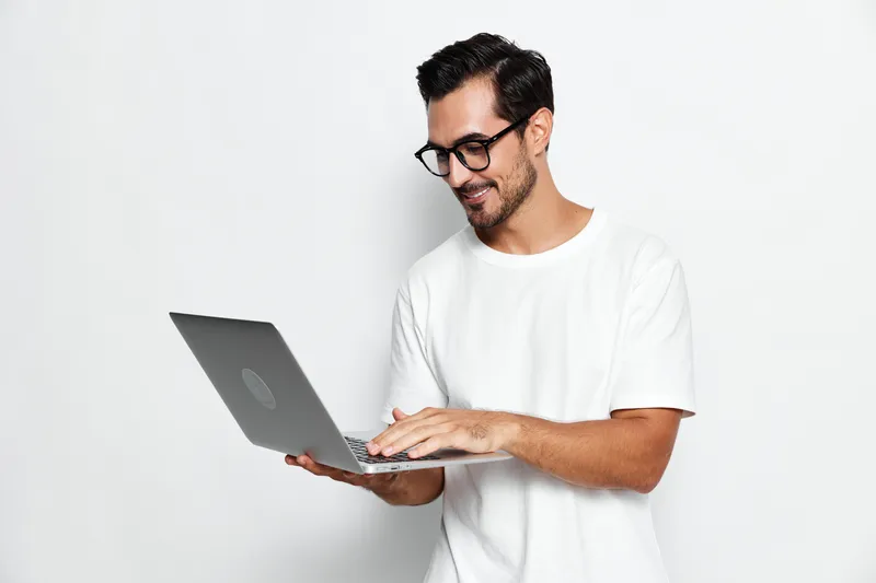 Smiling young man using laptop while standing against plain solid white background. Casual outfit with black glasses, showcasing modern technology and lifestyle.
