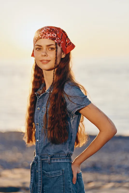 Young woman with long hair wearing red bandana and denim dress standing on the beach during golden hour with calm sea in the background.