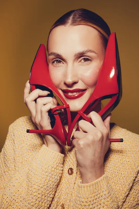 Smiling woman holds red high heels near face, wearing beige sweater. Studio portrait with warm lighting on brown background. Concept of fashion, style, and elegance.