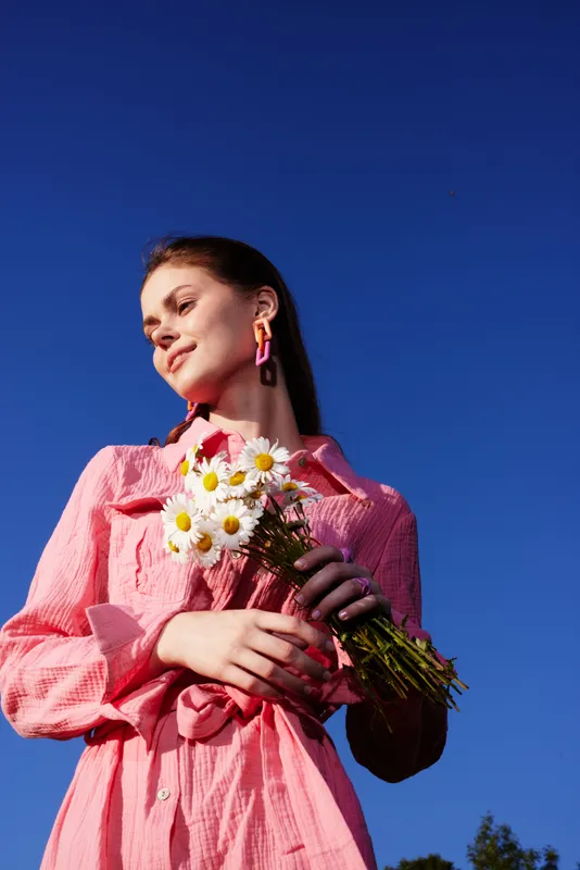 Young woman holding daisies, wearing a pink dress, against a clear blue sky, radiating joy and warmth in a vibrant summer setting