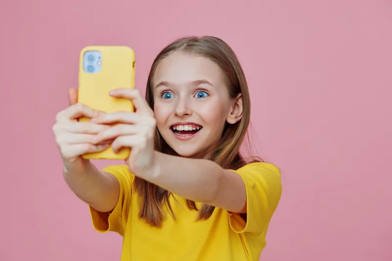 Happy girl taking a selfie with a smartphone against a pink background, wearing a yellow t shirt, expressing joy and excitement, showcasing youthful energy and vibrant colors