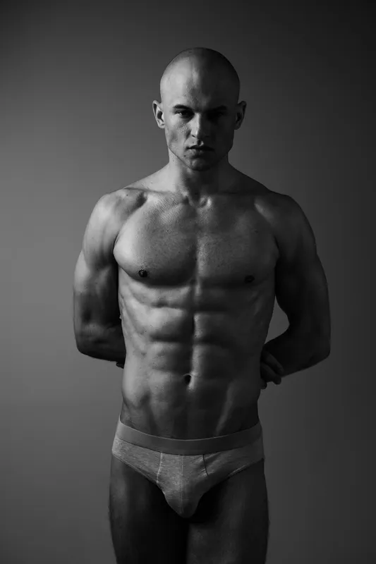 Muscular man posing confidently in black and white, showcasing strength and determination. The image emphasizes the male physique and powerful presence.