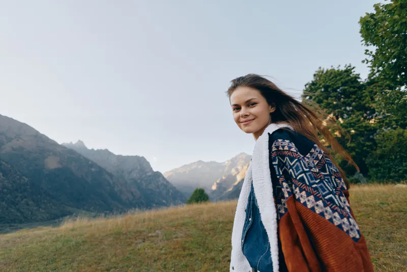 Woman smile mountains nature landscape sweater outdoors portrait: Young woman in patterned sweater walking on grassy hillside with a warm smile, looking back at scenic mountain vista in soft light.