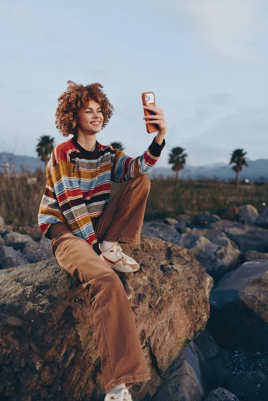 Woman lifestyle selfie outdoors, seated on rocks by shore, holding smartphone up, smiling with curly hair, casual sweater and pants, warm sunset sky, travel mood, natural light