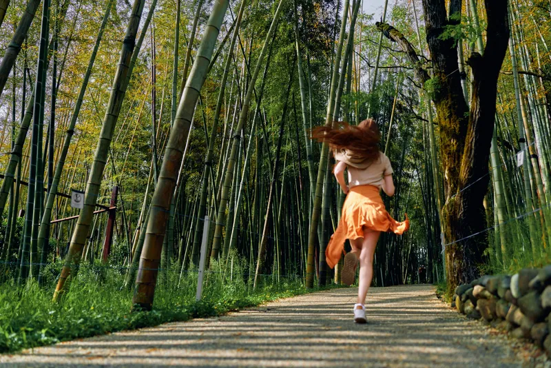 Woman running through bamboo forest on a sunlit path, orange skirt flowing and hair in motion, outdoor nature scene with dynamic movement and serenity