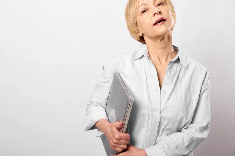 Elderly woman holding a book, neck pain expression, indoor portrait on white background, concerned mood, soft natural light, calm stance, with copy space