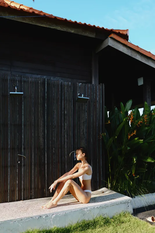 Relaxed woman in bikini sits on a concrete ledge beside an outdoor shower, tropical plants in the background, basking in sun, serene moment of self care and summer lifestyle.
