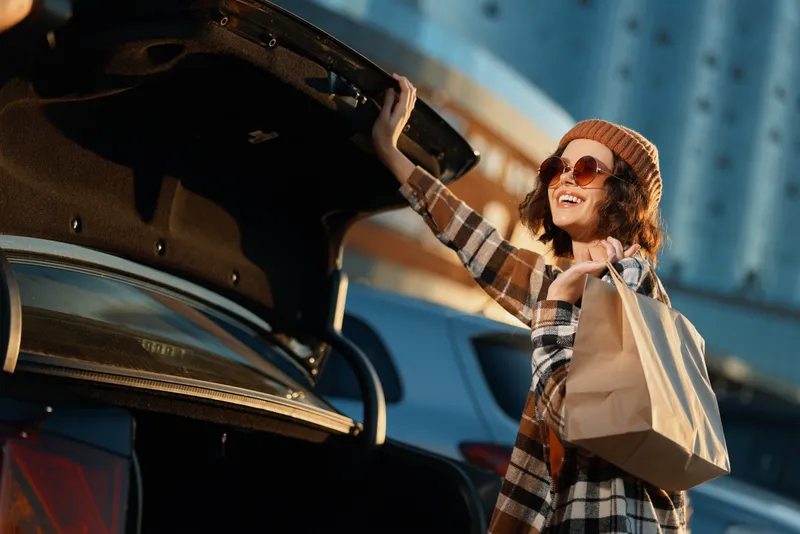 Woman shopping at car trunk, smile with paper bag in hand in warm sunlight. Urban lifestyle portrait with golden hour glow, authenticity, candid lifestyle, mindful living and emotional storytelling.
