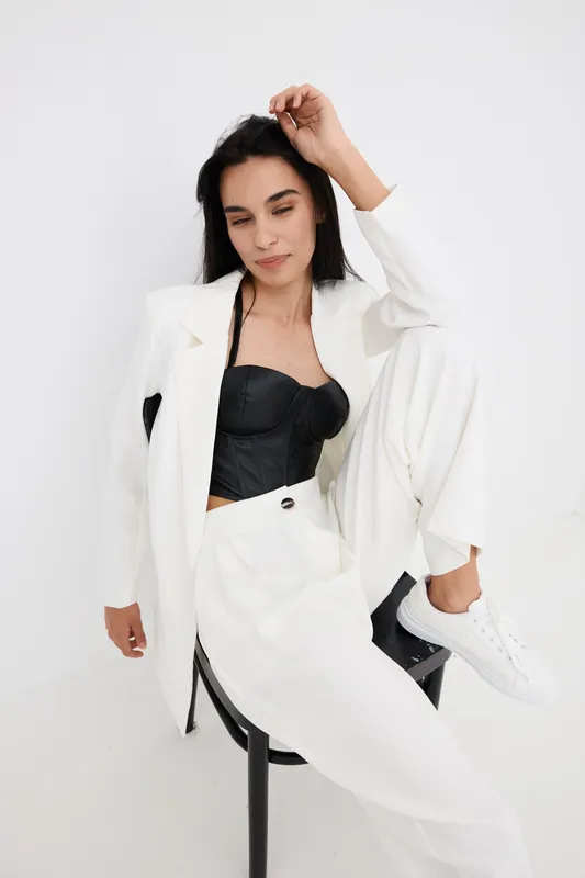 stylish woman in white suit and black top sitting relaxed on chair with casual white sneakers, minimalistic studio background, modern fashion look