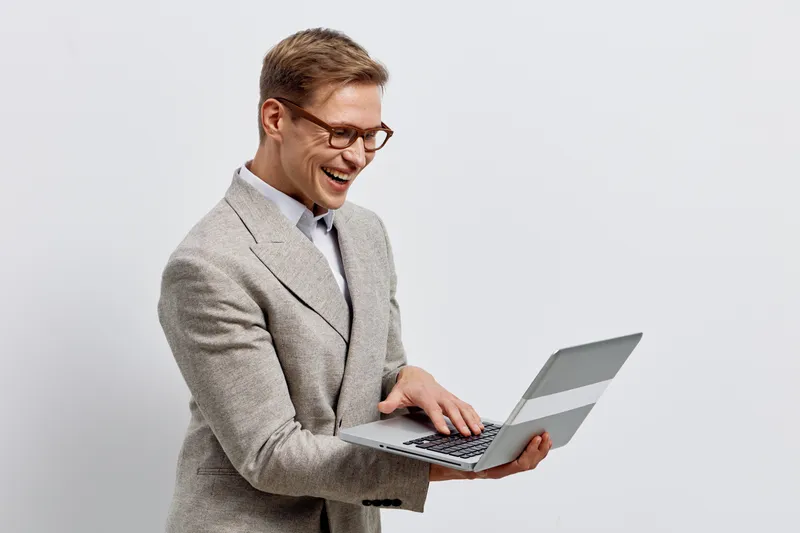 Professional smiling man in smart casual attire holding a laptop computer in hand, looking at the screen, isolated on plain white background. Business, technology, communication concept.