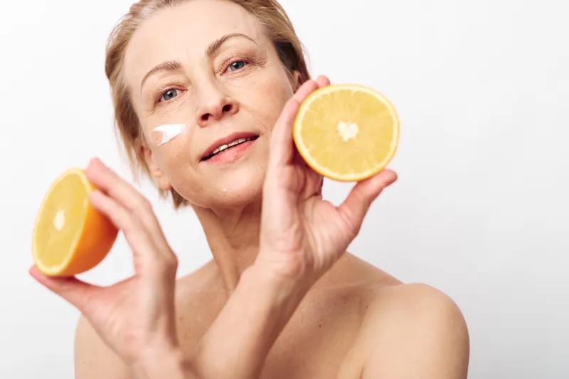Mature woman with natural skin applying cream holds two halves of fresh orange fruit close to face isolated on white. Concept of skincare, vitamin c, healthy lifestyle and beauty care.