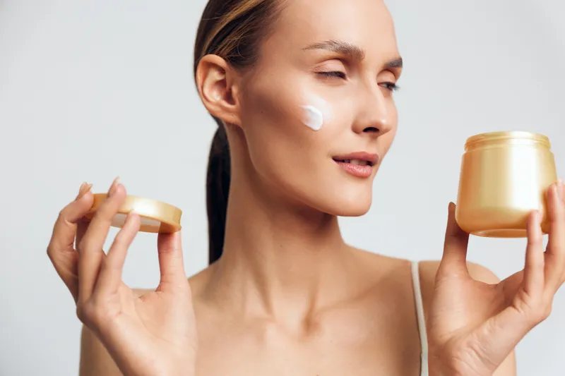 Young woman with clear skin holding an open jar of cream applying moisturizer on her cheek. Skincare and beauty concept with soft natural lighting on white background.
