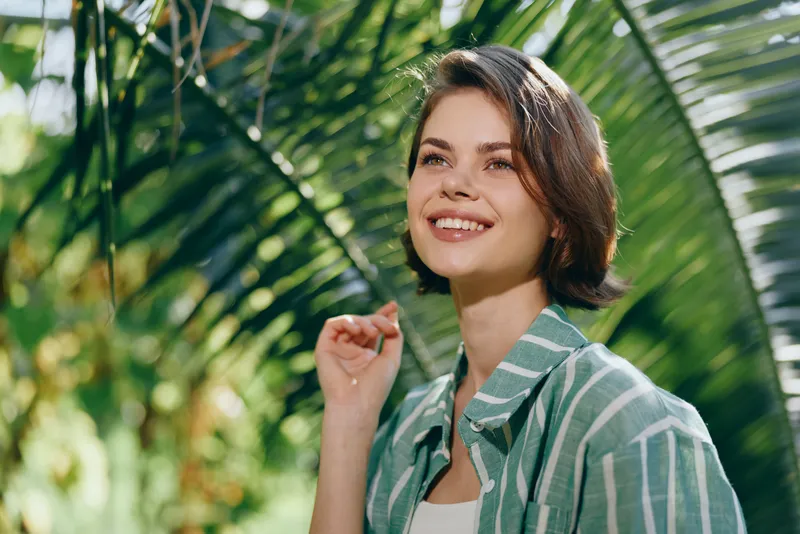 A smiling woman stands among lush tropical plants, bathed in natural light, wearing a striped shirt.