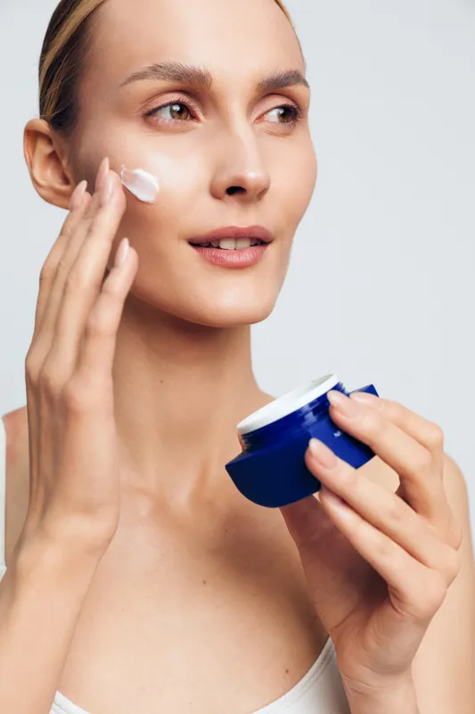 Woman applying skincare cream to face, holding cosmetic jar near cheek, clean studio background, calm expression, beauty treatment, spa hygiene, soft light gently