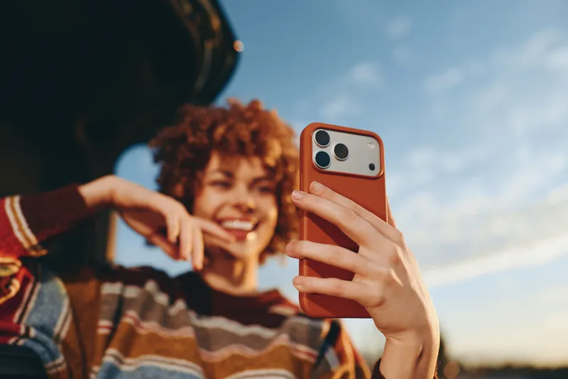 Woman selfie smartphone curly hair smiling outdoor lifestyle, foreground hand holds phone with brown case, background woman grins, curly hair and striped sweater, blue sky bokeh, daylight