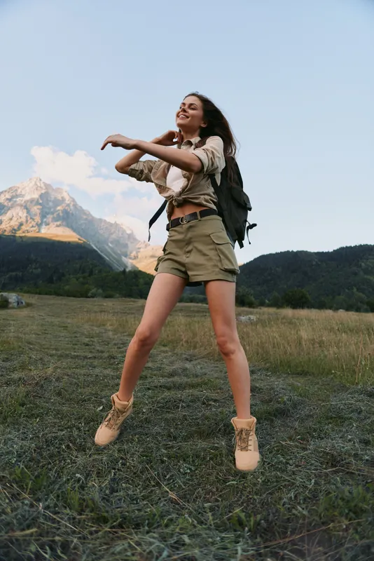 A young woman leaps across a grassy field with a backpack, set against a vast mountain landscape, conveying carefree outdoor adventure, vitality, and a bold sense of freedom.