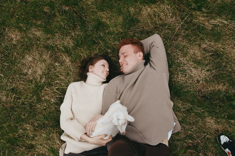 A cozy couple lies on grass in knit sweaters, smiling and cuddling a white goat kid between them. A warm, natural lifestyle moment for romance, companionship, and rural animal friendship outdoors,
