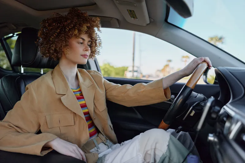 A woman in retro style drives a car, wearing a tan jacket and striped tee, focused on the road. A candid lifestyle moment with warm film tones and relaxed, urban mood.