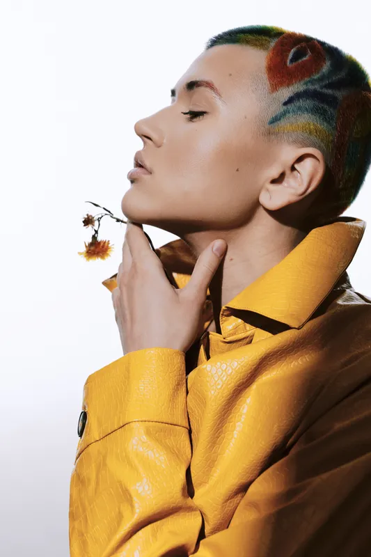 Young woman with colorful artistic haircut in yellow jacket holds dried flower near face, showing delicate expression and modern style with bright contrast lighting.