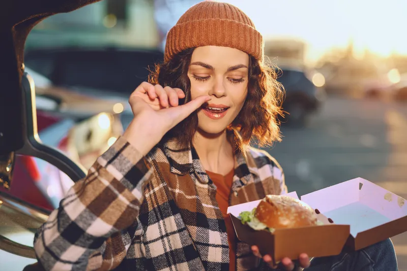 Woman holding burger outdoors by a car trunk at golden hour, candid smile and beanie with plaid jacket, authenticity and candid lifestyle vibe, emotional storytelling and mindful living.