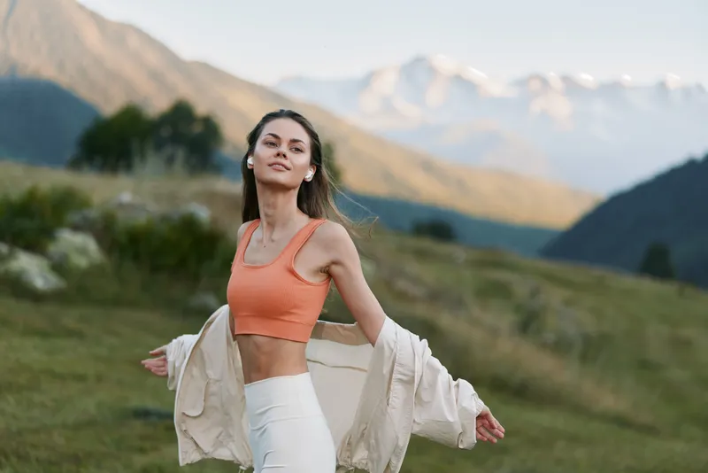 A cheerful woman wearing an orange sports top stands outdoors in a scenic valley with mountains in the background, smiling as she embraces the sunshine and fresh air.