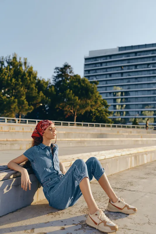 Lifestyle woman in modern boho-western denim street style sits relaxed outdoors with film color tones, casual outfit, red bandana, and comfortable sneakers under clear sky.