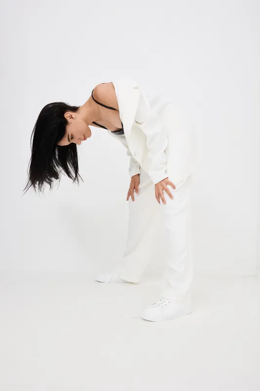 Woman bending forward in loose white suit with black innerwear and white sneakers against white background, showcasing casual contemporary fashion and relaxed posture.