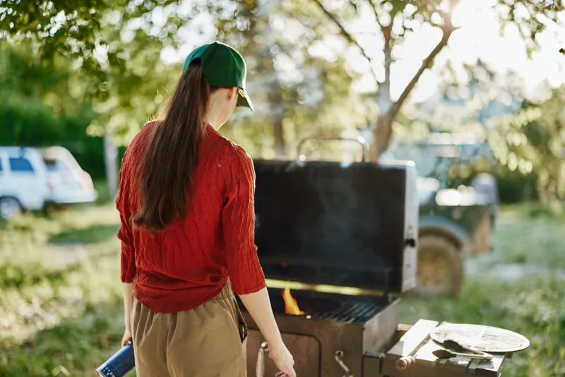 Young woman grilling outdoors in nature, wearing casual clothing, enjoying a sunny day, with greenery in the background and a barbecue setup