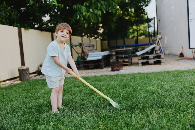 A cheerful boy with short hair digging in the garden with a yellow shovel, enjoying a sunny day outdoors, surrounded by green grass and wooden pallets.