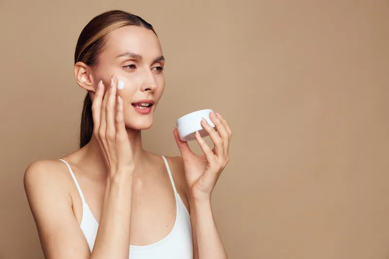 Young woman applies face cream on clean skin holding jar with moisturizer isolated on beige background with natural skincare beauty routine concept.