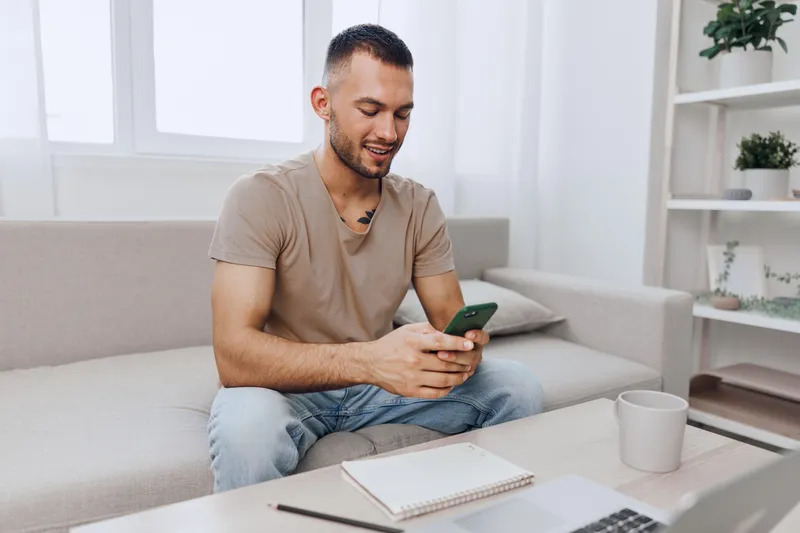 Happy man using smartphone while sitting on cozy couch, dressed casually in a light t-shirt and jeans. Bright, modern interior with natural light enhances the atmosphere.