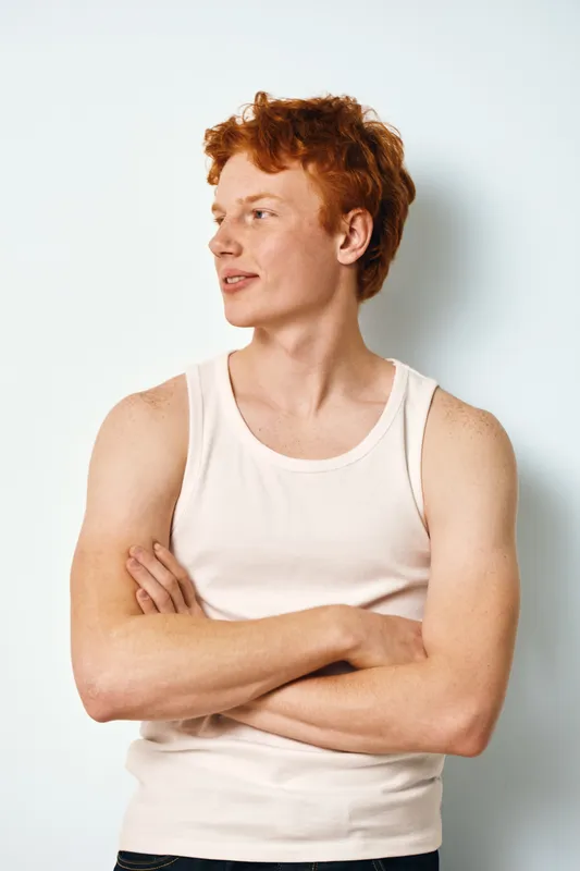 Young man with red hair wearing white tank top posing with arms crossed looking sideways against white background. Casual style and confident expression in studio portrait.