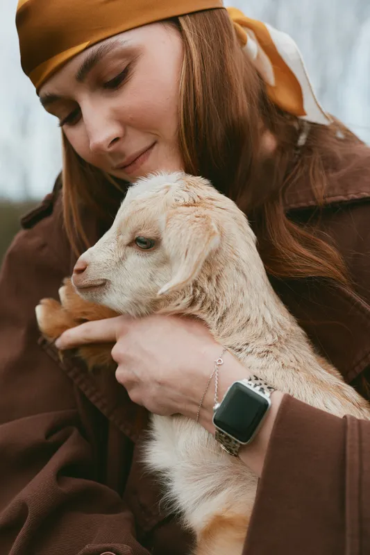 Lifestyle moment of a woman outdoors with a gentle goat kid, close cuddle, warm tones, light breeze, smiling expression, rustic clothing, natural setting, calm bond with animal, serene countryside
