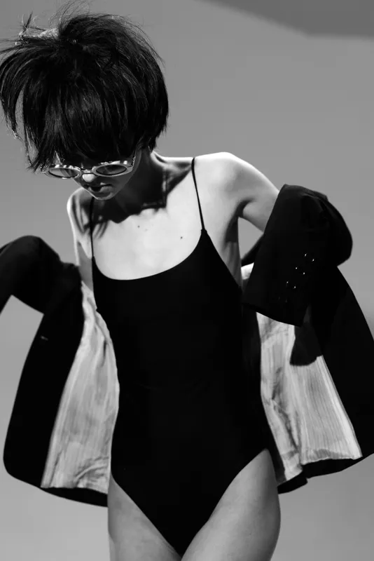 Black and white image of a young skinny woman wearing sunglasses and a black bodysuit, putting on a jacket with stylish short hair.