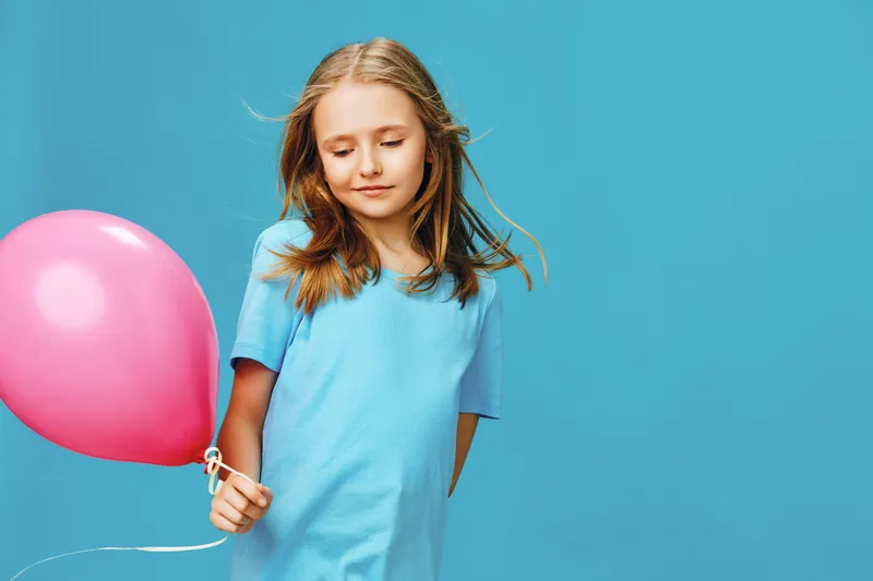 A cheerful young girl holding a pink balloon against a bright blue background, expressing joy and innocence in a playful and vibrant setting
