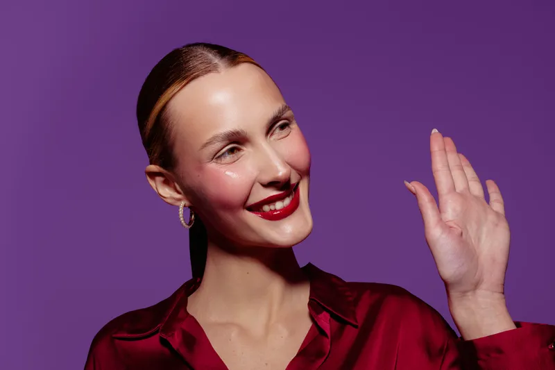Smiling woman with red lipstick and earrings waves hand on purple background. Studio portrait of happy female model in red blouse with natural makeup and slicked hair.