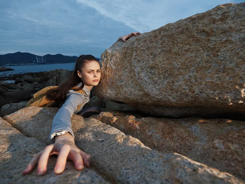 A determined young woman climbing a rocky cliff, showcasing strength and resilience against a dramatic coastal backdrop during sunset