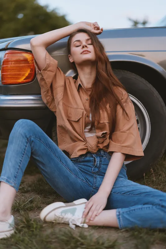 Young woman in casual fashion poses next to a vintage car, showcasing a relaxed vibe and summer style with denim jeans and a brown shirt.