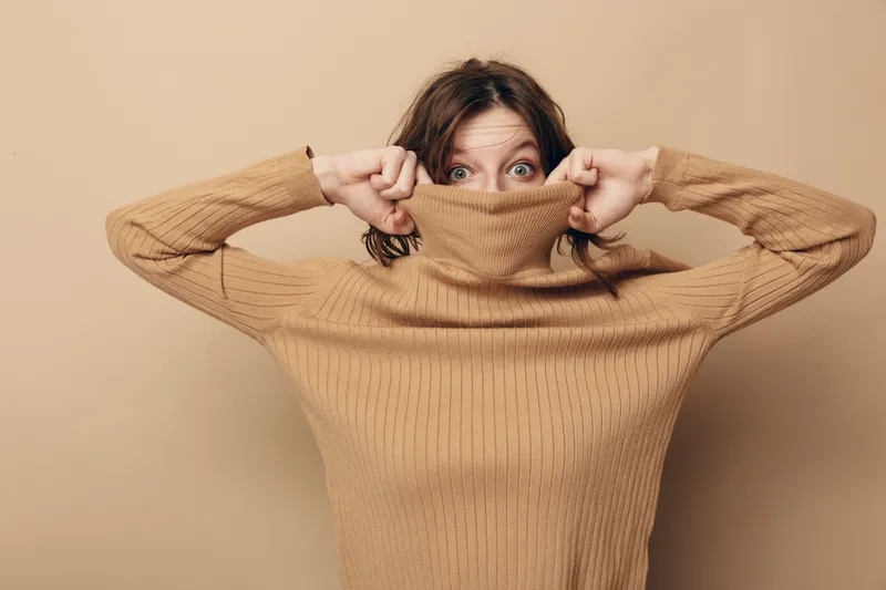Young woman with blue eyes wearing beige knitted sweater pulls collar over her face, looking surprised. Neutral background, casual style, cozy autumn or winter fashion concept.