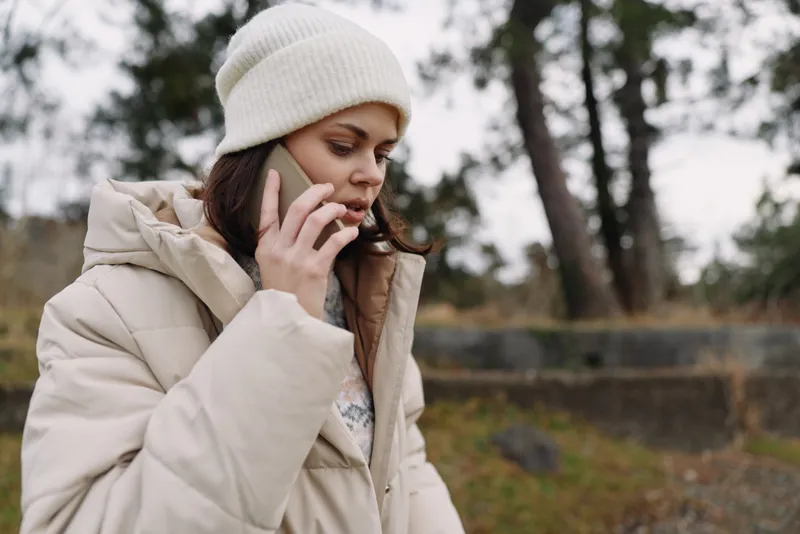 Woman on outdoor winter scene talking on phone, wearing a jacket and beanie in a park, cold weather mood, candid portrait showcasing casual lifestyle in nature.