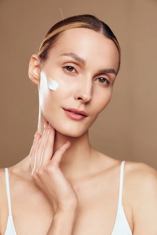 Young woman with clean skin applies moisturizing cream on face with hand isolated on beige background natural beauty healthy skincare routine closeup portrait