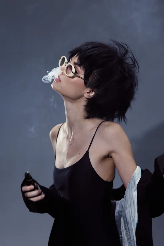 Stylish woman with sunglasses exhaling smoke wearing black clothing against dark background
