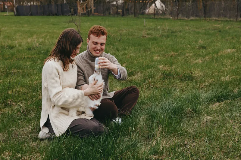 A young couple sits on green grass in casual sweaters enjoying a playful moment with a white baby goat in a relaxed outdoor lifestyle setting during early spring.